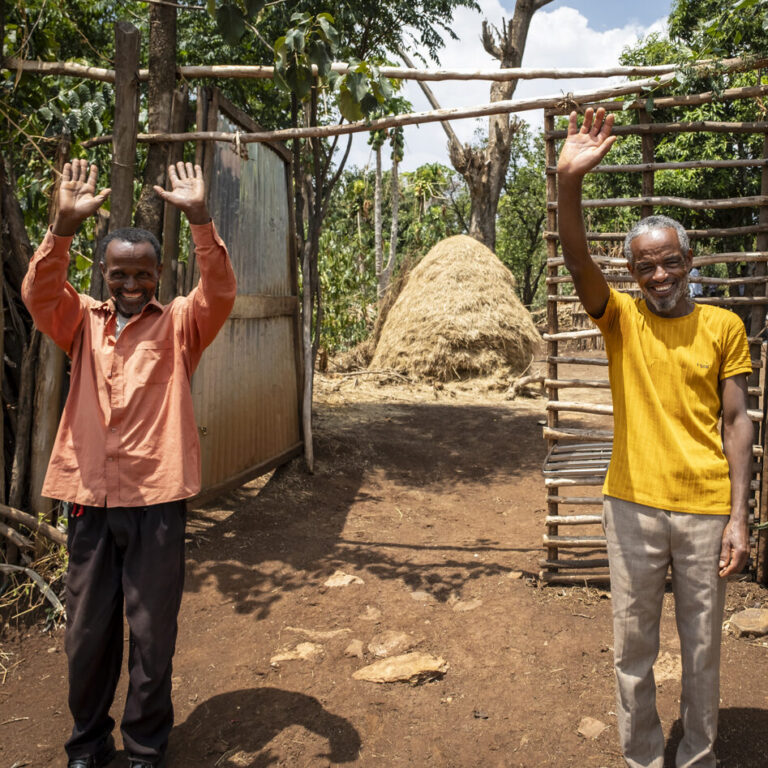 Two men are standing outside by a haystack and some trees, smiling and waving, looking friendly and welcoming.