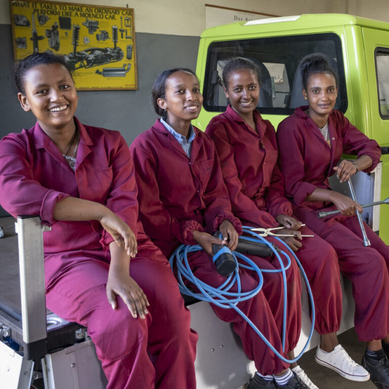 Four women in maroon overalls smile and hold tools and cables in a car repair shop.