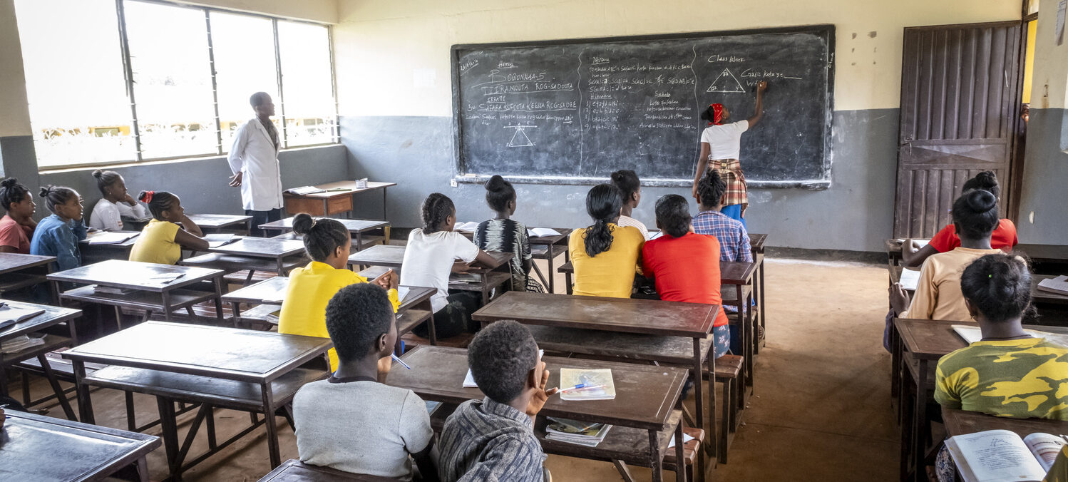 Students at their desks observe a girl writing on a blackboard in the classroom and presenting educational initiatives.