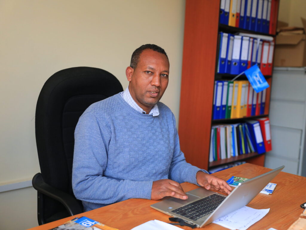 Man in grey jumper with laptop at office desk with laptop and colourful files in the background.