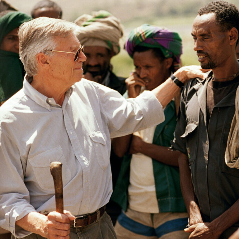 An elderly man wearing glasses converses with a younger man in an outdoor group and suggests community support.