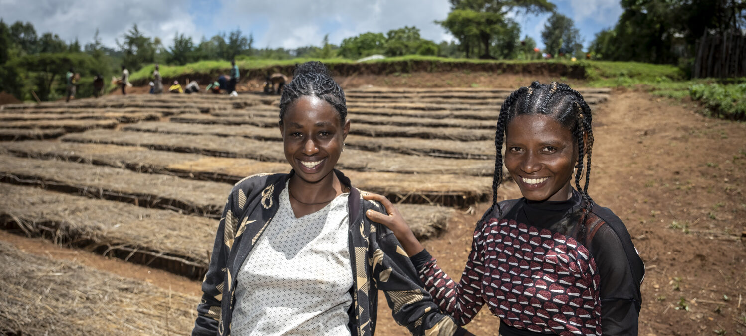 Two women standing smiling next to the sun-dried beds of a charity-funded agricultural project.