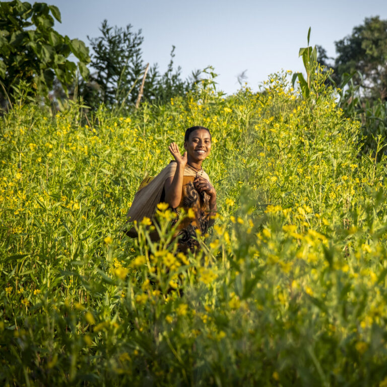 A woman is walking across a field with yellow flowers. She carries a water canister on her back.