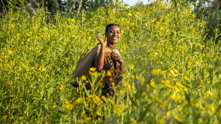 A woman is walking across a field with yellow flowers. She carries a water canister on her back.