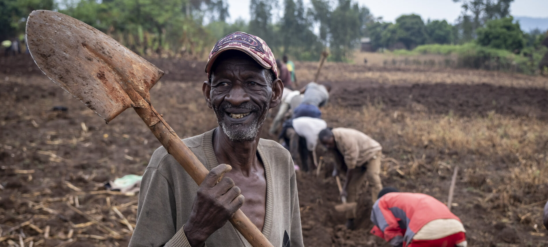 A man is standing on a field with a shovel. In the background, several other men are working.