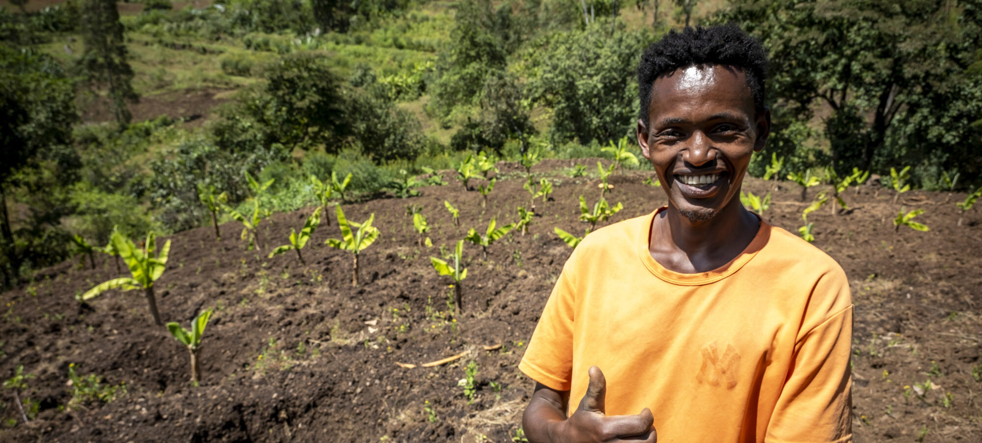 Ein junger Mann blickt lächelnd in die Kamera. Er steht vor einem Feld mit Pflanzen.