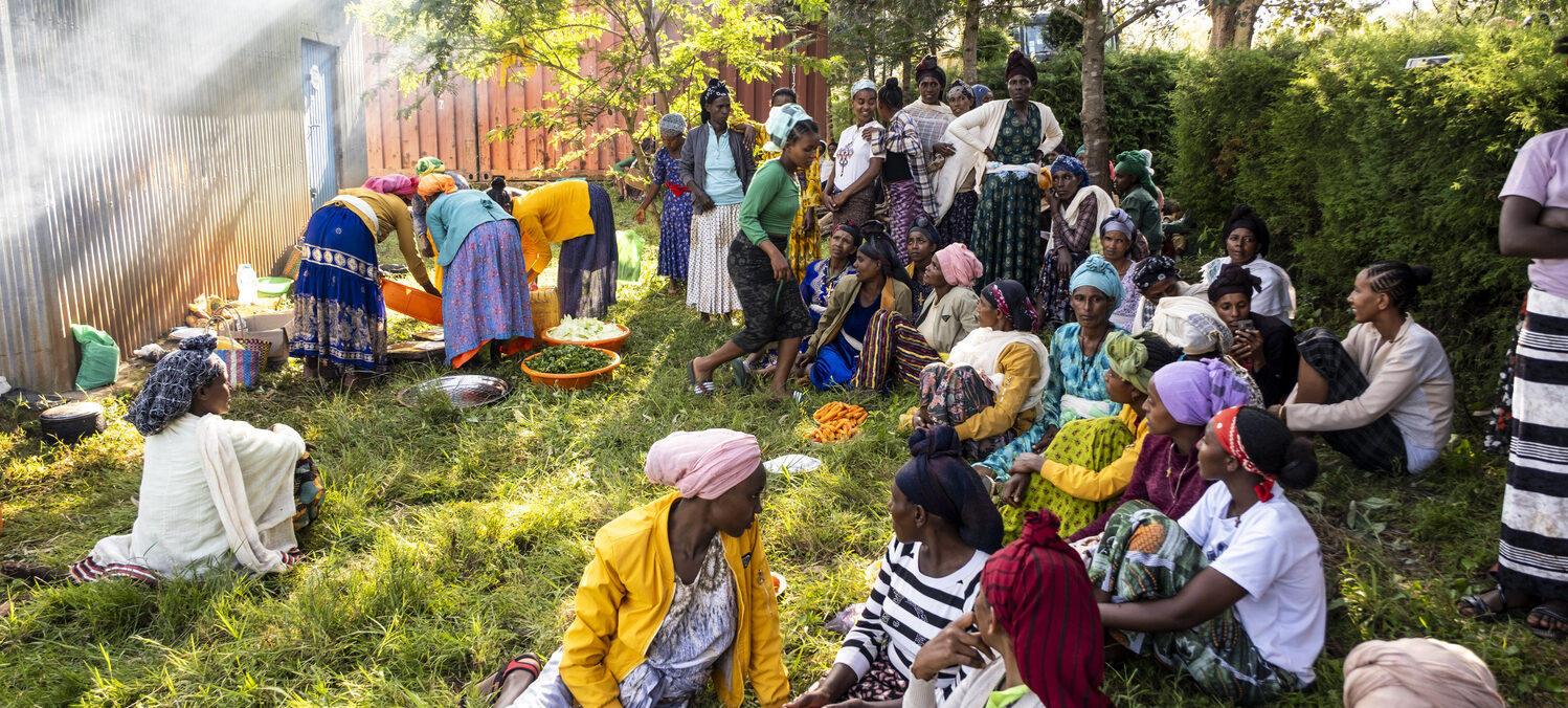 A group of women wearing colorful dresses are sitting outdoors in the greenery and talking.