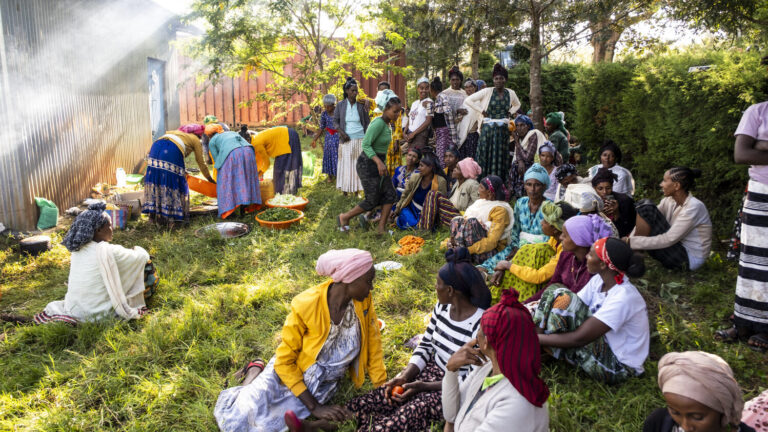 A group of women wearing colorful dresses are sitting outdoors in the greenery and talking.