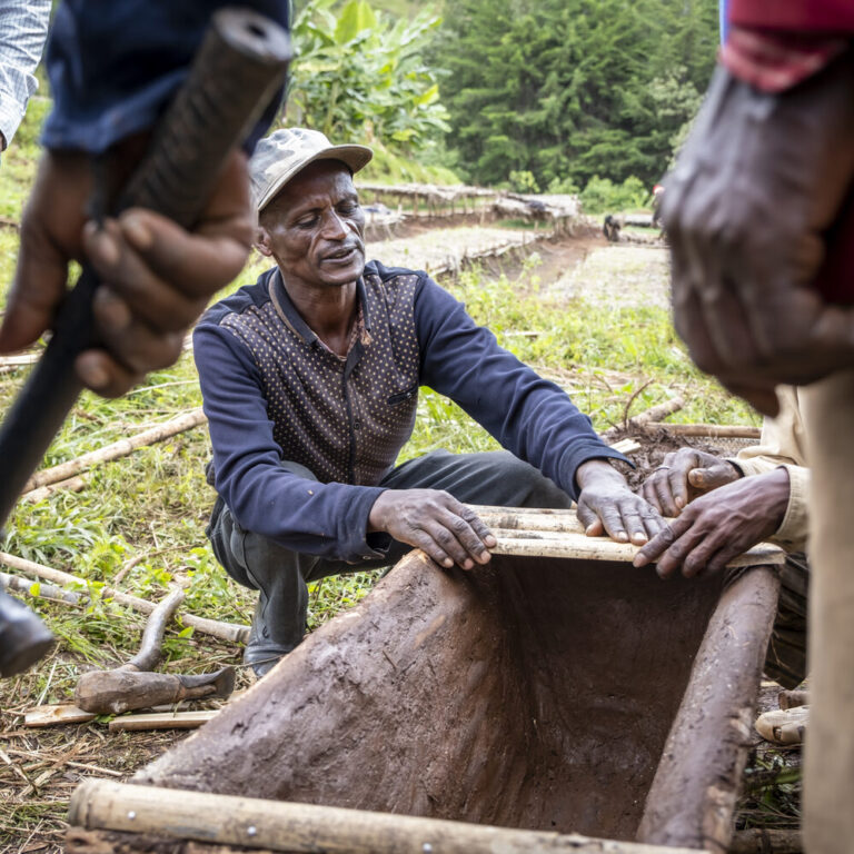 A man kneels outdoors and builds a wooden structure while several people stand around him.