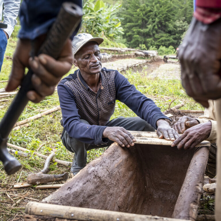A man kneels outdoors and builds a wooden structure while several people stand around him.