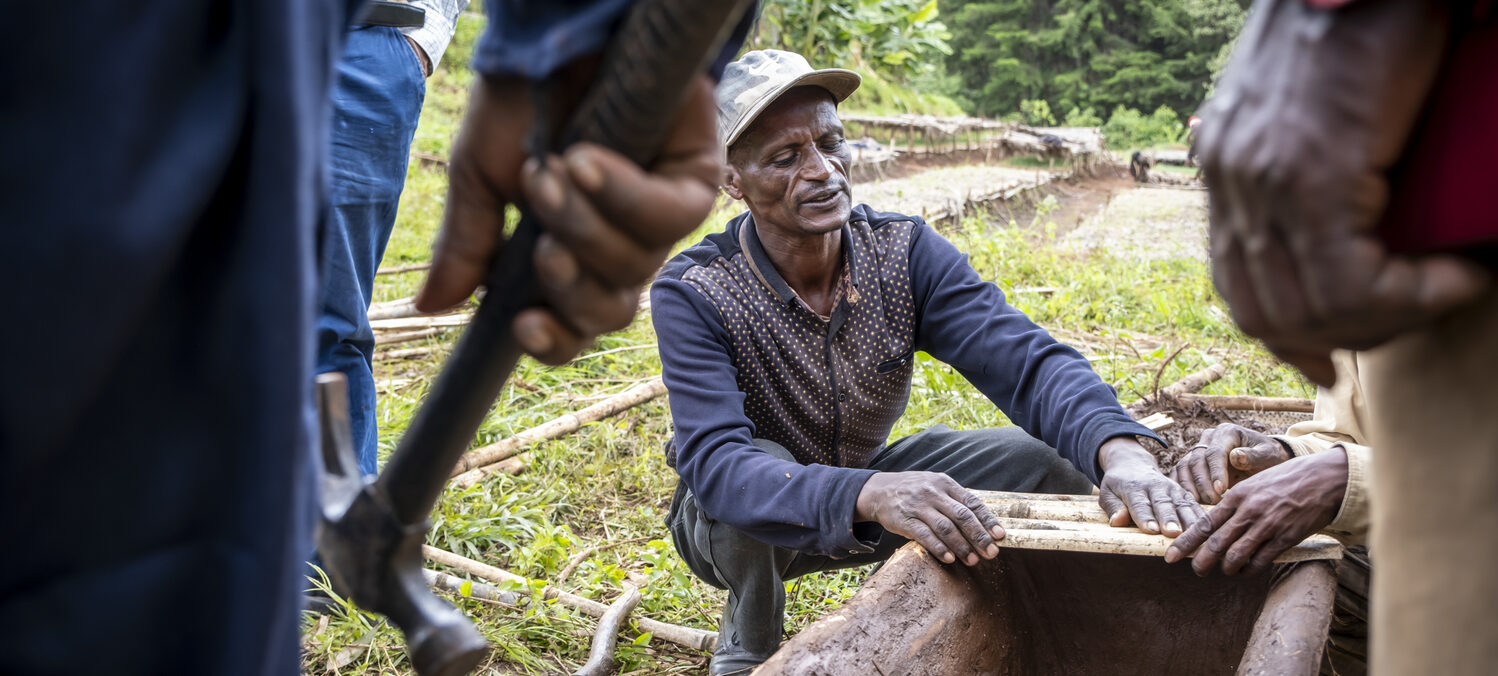 A man kneels outdoors and builds a wooden structure while several people stand around him.