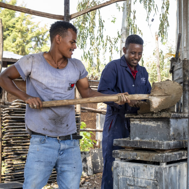 Two men are shoveling wet mud into a brick-making machine outdoors.