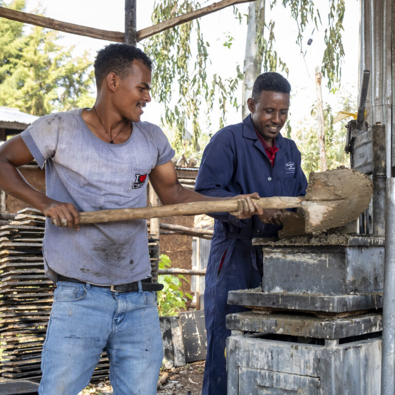 Two men are shoveling wet mud into a brick-making machine outdoors.