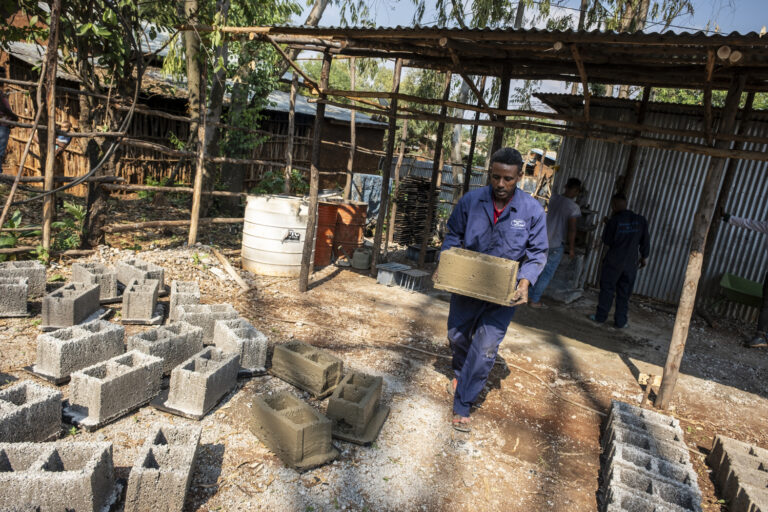 A man carries a concrete block in a sunny outdoor workshop.