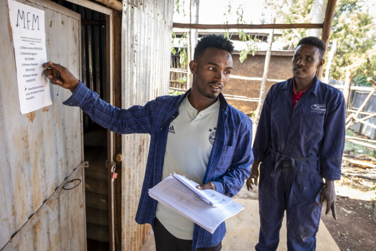 Two men stand in front of a building, checking the papers on the door.