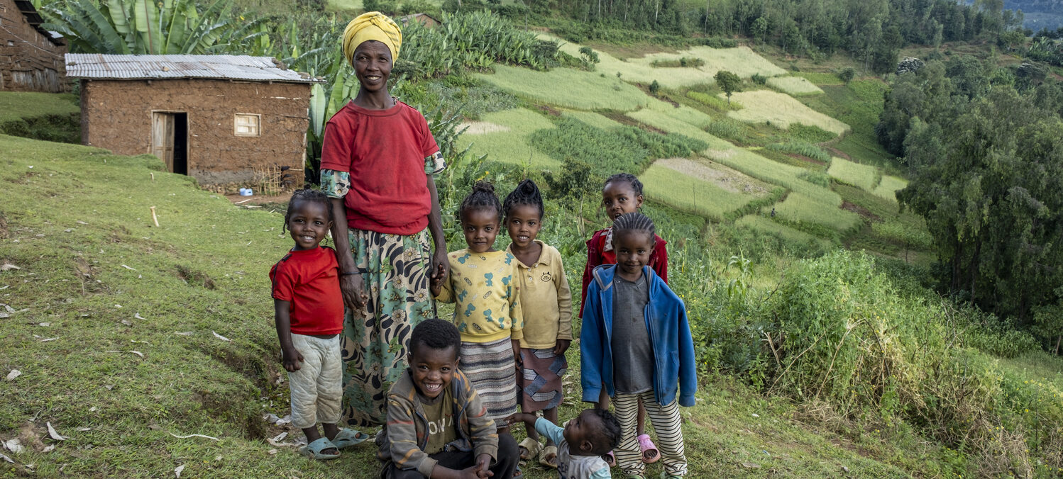 A woman with seven children stands on a grassy hill; fields and a house can be seen behind them.