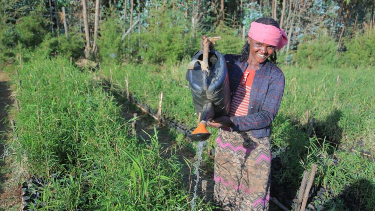 A woman smiling while watering plants with a watering can.
