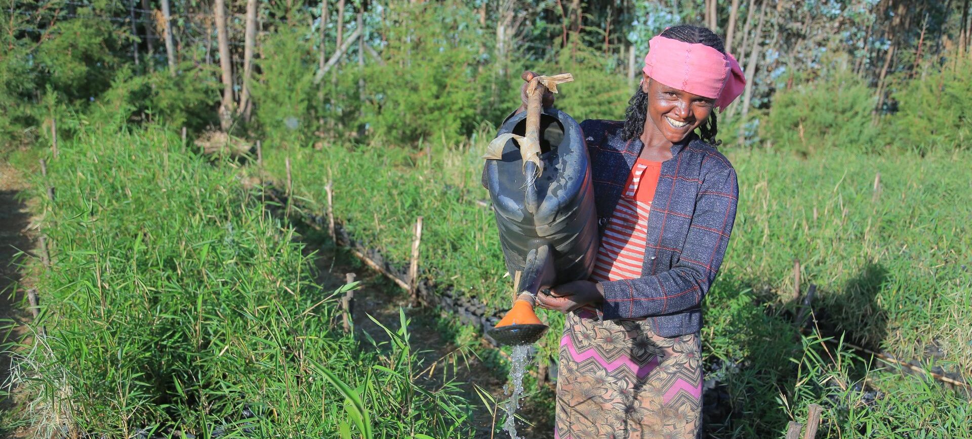 A woman smiling while watering plants with a watering can.
