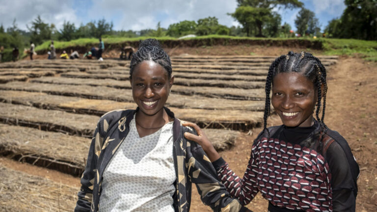 Two women standing smiling next to the sun-dried beds of a charity-funded agricultural project.