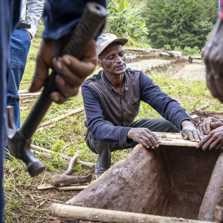 A man kneels outdoors and builds a wooden structure while several people stand around him.