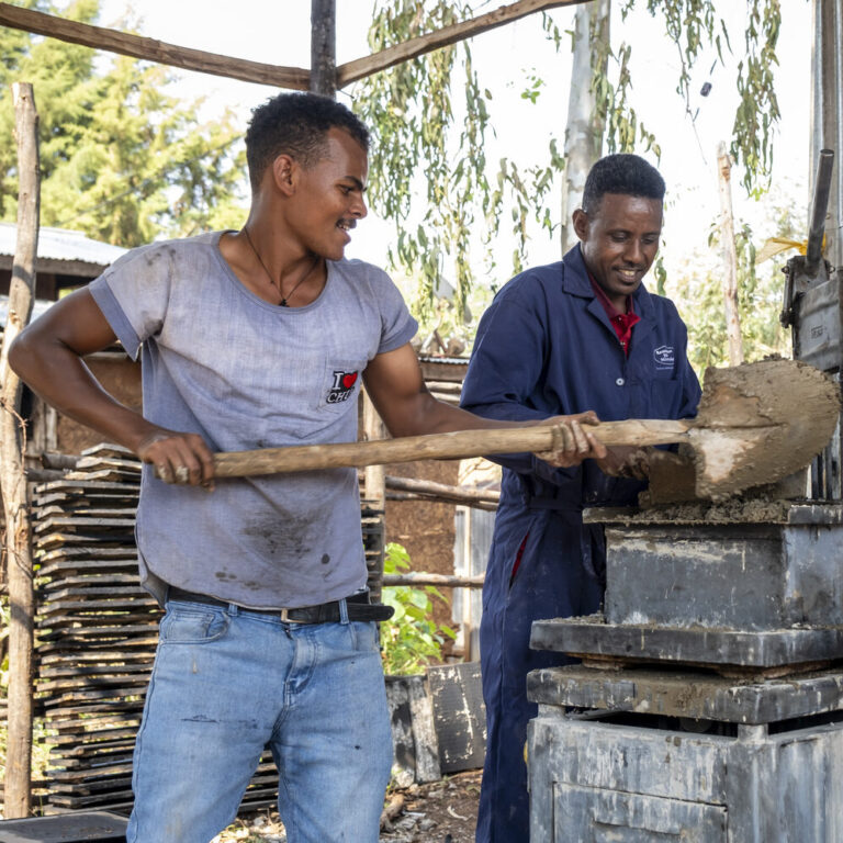 Two men are shoveling wet mud into a brick-making machine outdoors.