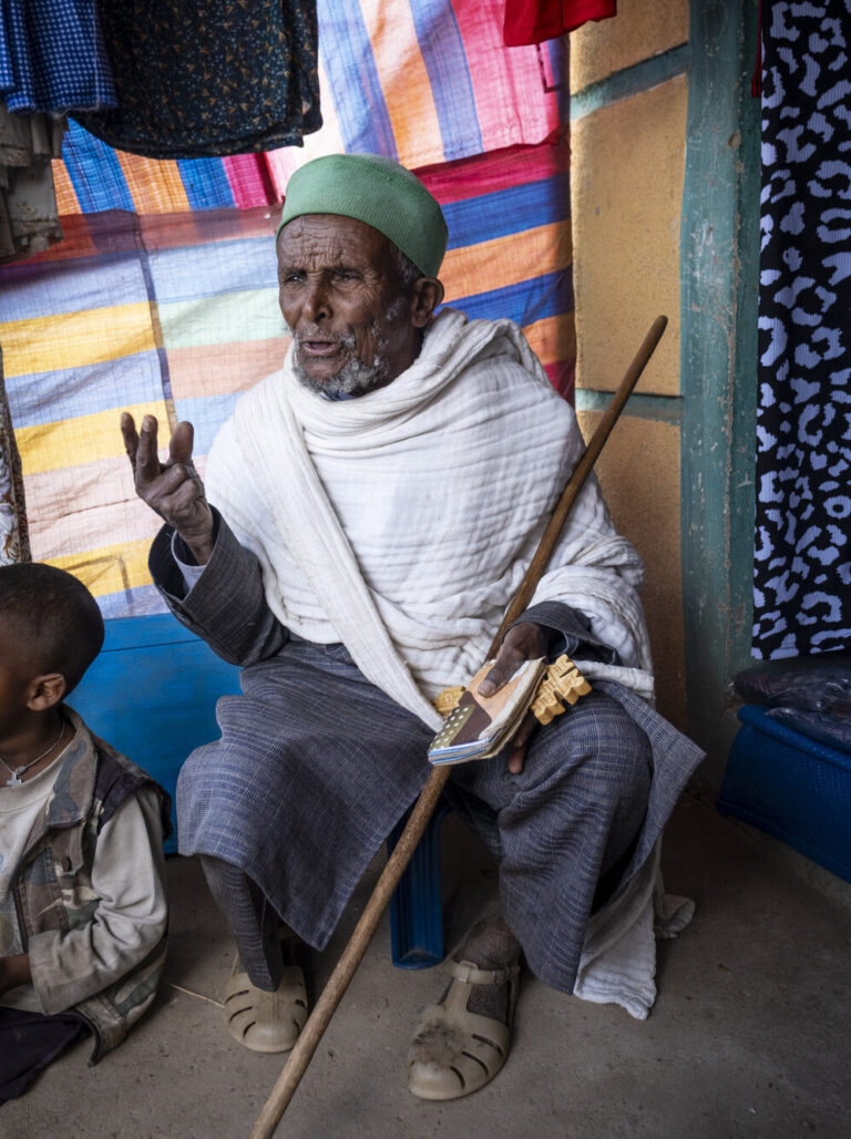 An old man holding a book and a stick while sitting and speaking.